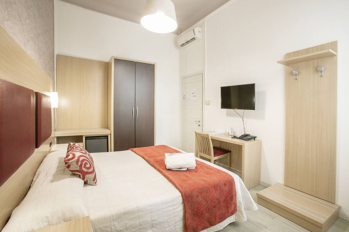 A hotel room with a bed, a desk, a chair, a TV, and a wardrobe.
