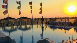 Una piscina con numerose bandiere, circondata da piante in vaso e un tramonto sullo sfondo.
