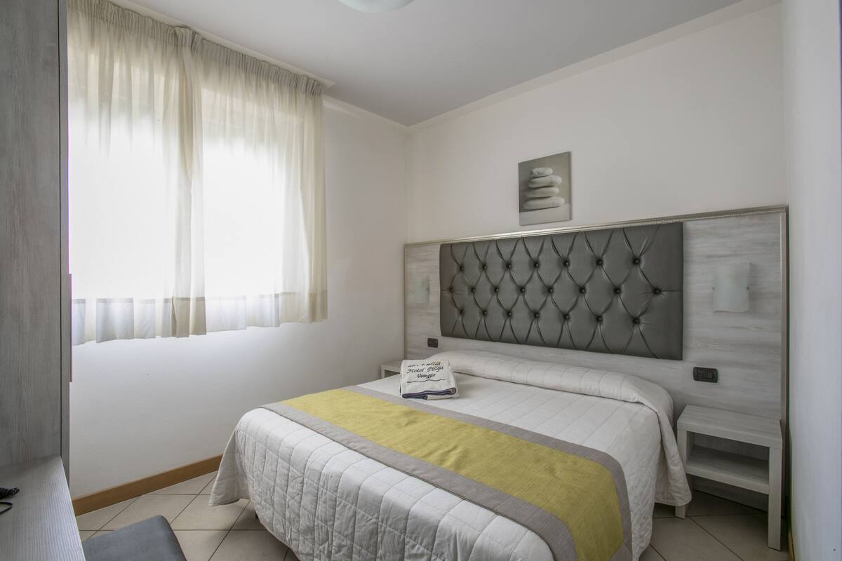A neatly arranged bedroom with a large bed, a bedside table, a nightstand, a window with curtains, and a wall-mounted artwork.