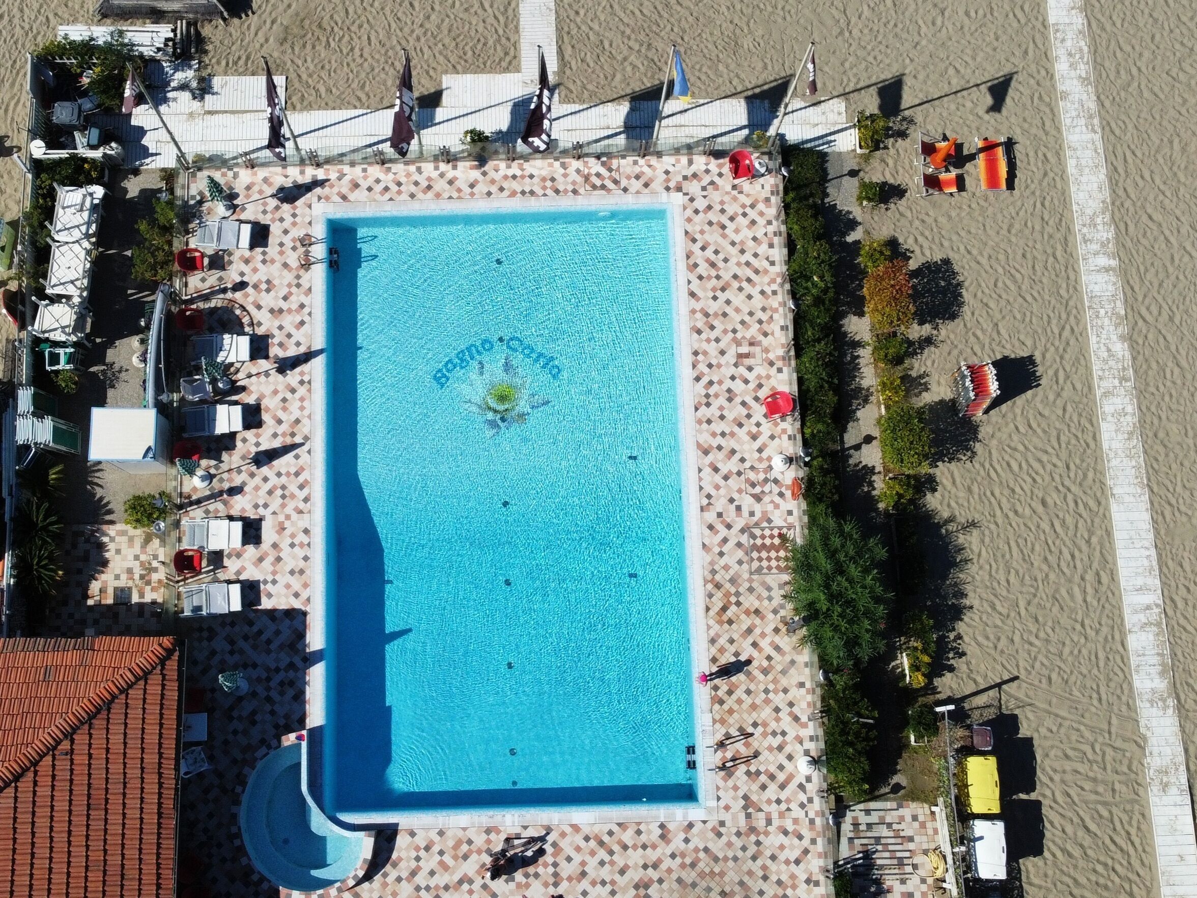 An aerial view of a rectangular swimming pool with a tiled deck surrounding it, a small circular fountain in the center, and a beach area with umbrellas and lounge chairs.
