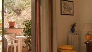 A room with a view of a hillside, a white chair, a table, a potted plant, a radiator, a lamp, and a framed picture on the wall.