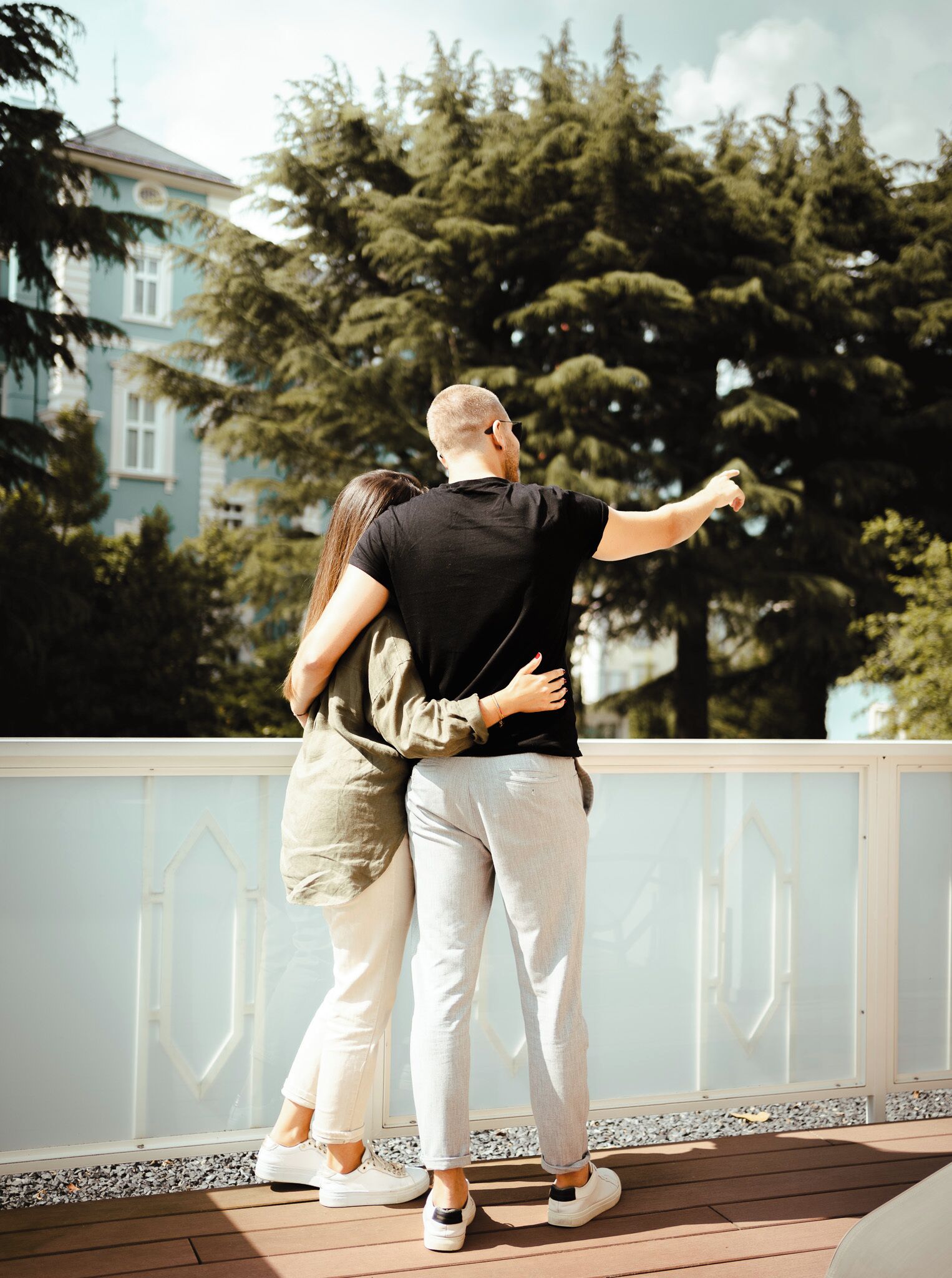 A couple embracing on a wooden deck with a railing, overlooking a building and trees.