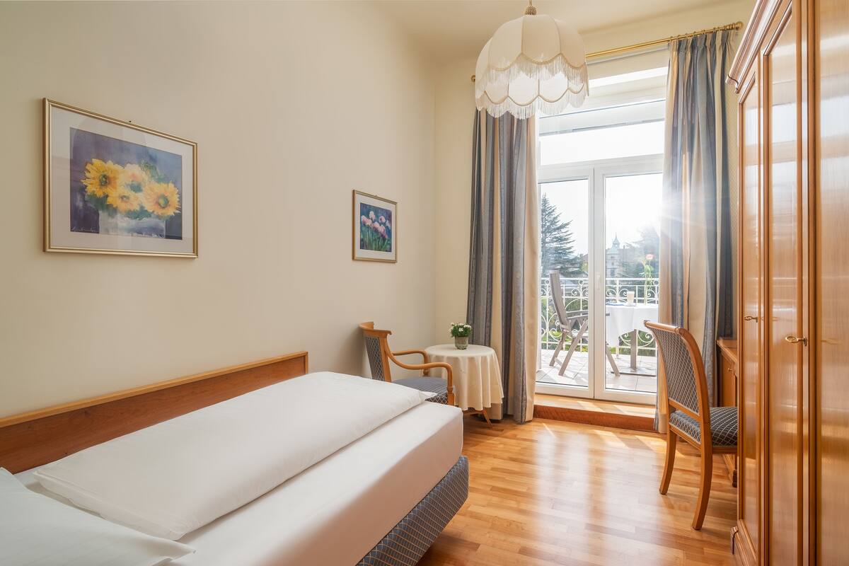 A hotel room with a bed, a chair, a small table with a plant, and a view of the outside through a glass door.