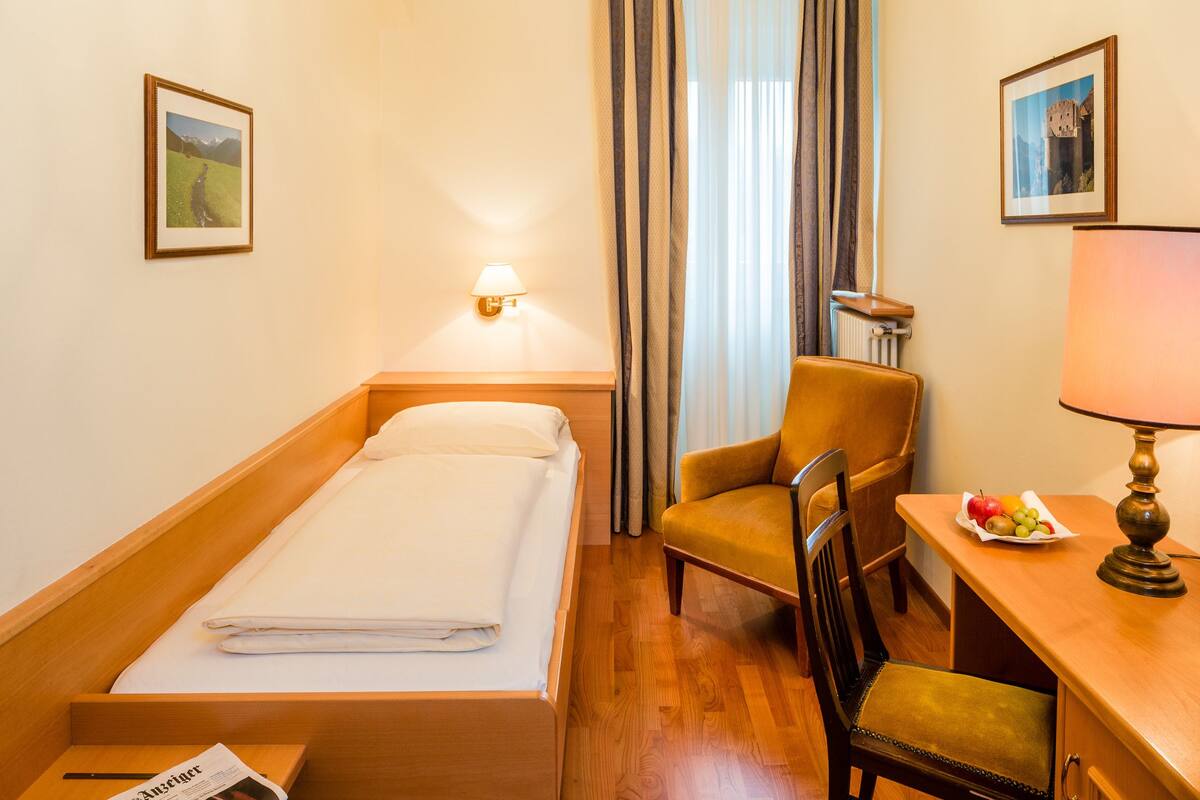 A hotel room with a bed, a chair, a desk with a lamp, and a table with fruit.