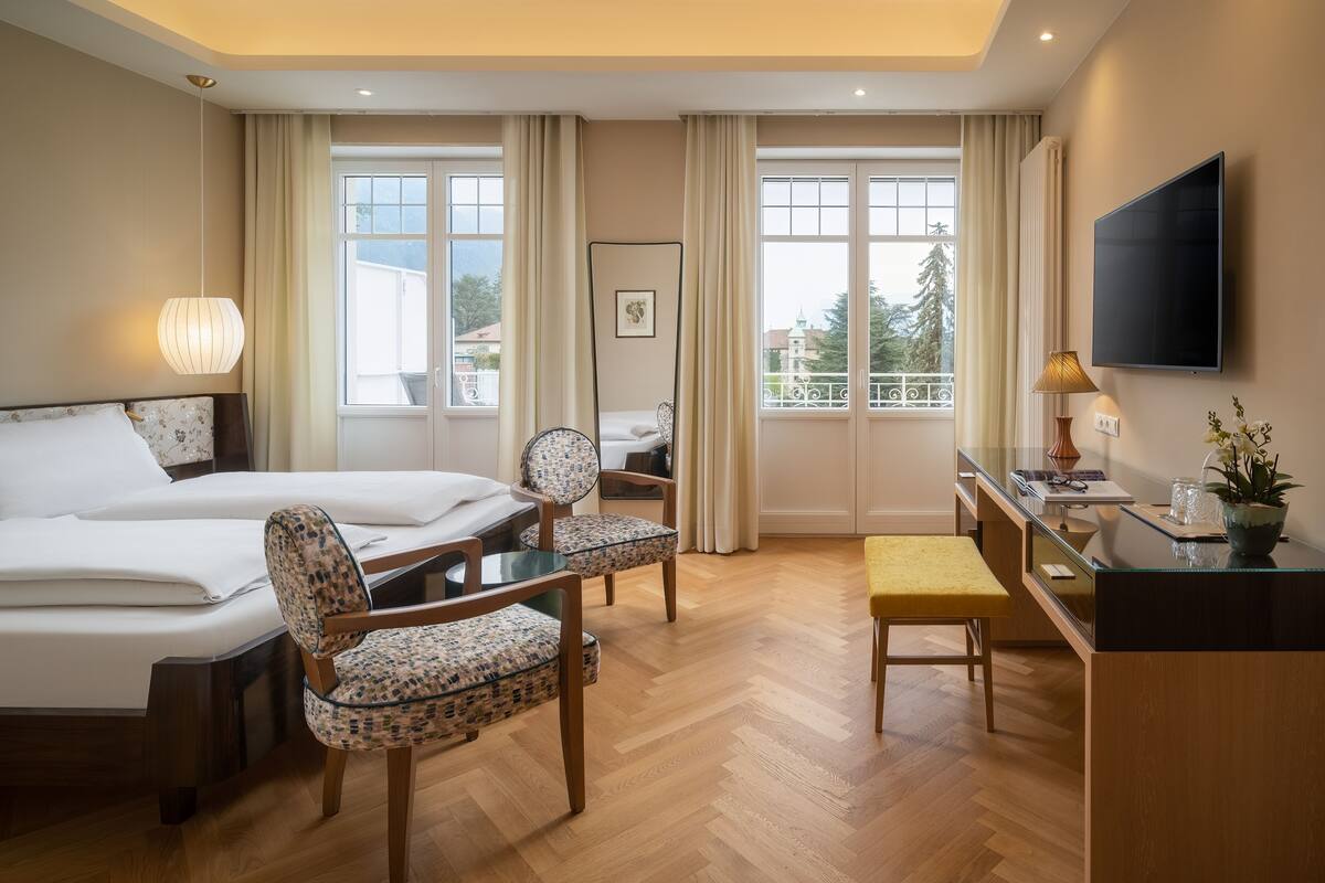 A hotel room with two beds, a desk, a chair, a TV, and large windows with curtains.