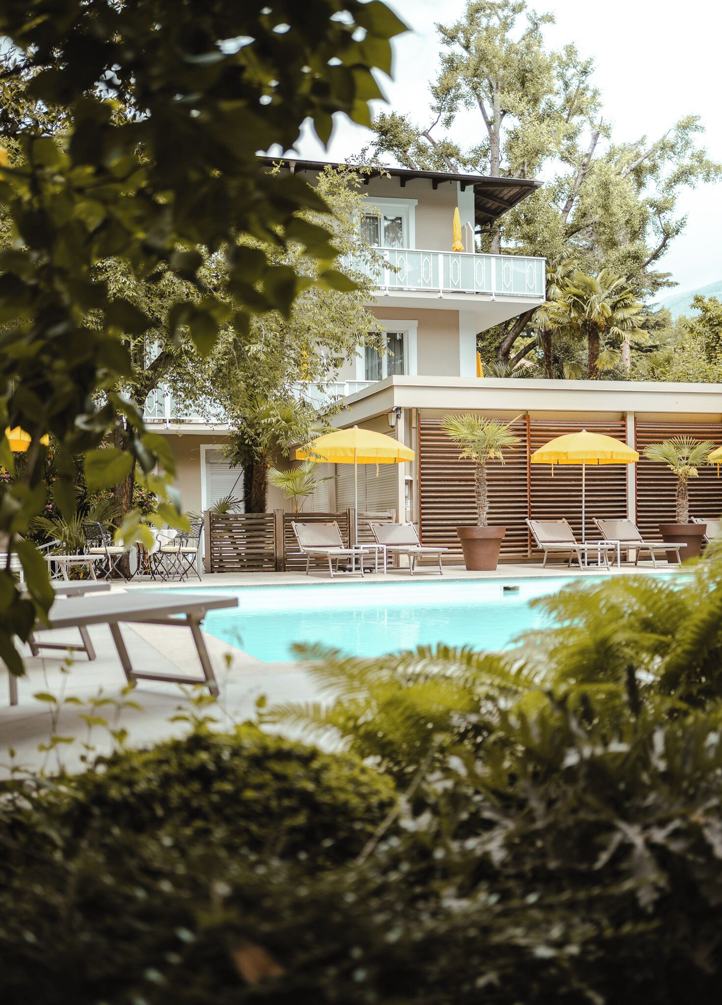 Un espace piscine agrémenté de chaises longues et de parasols, entouré de verdure et bordé d’un bâtiment doté d’un balcon.