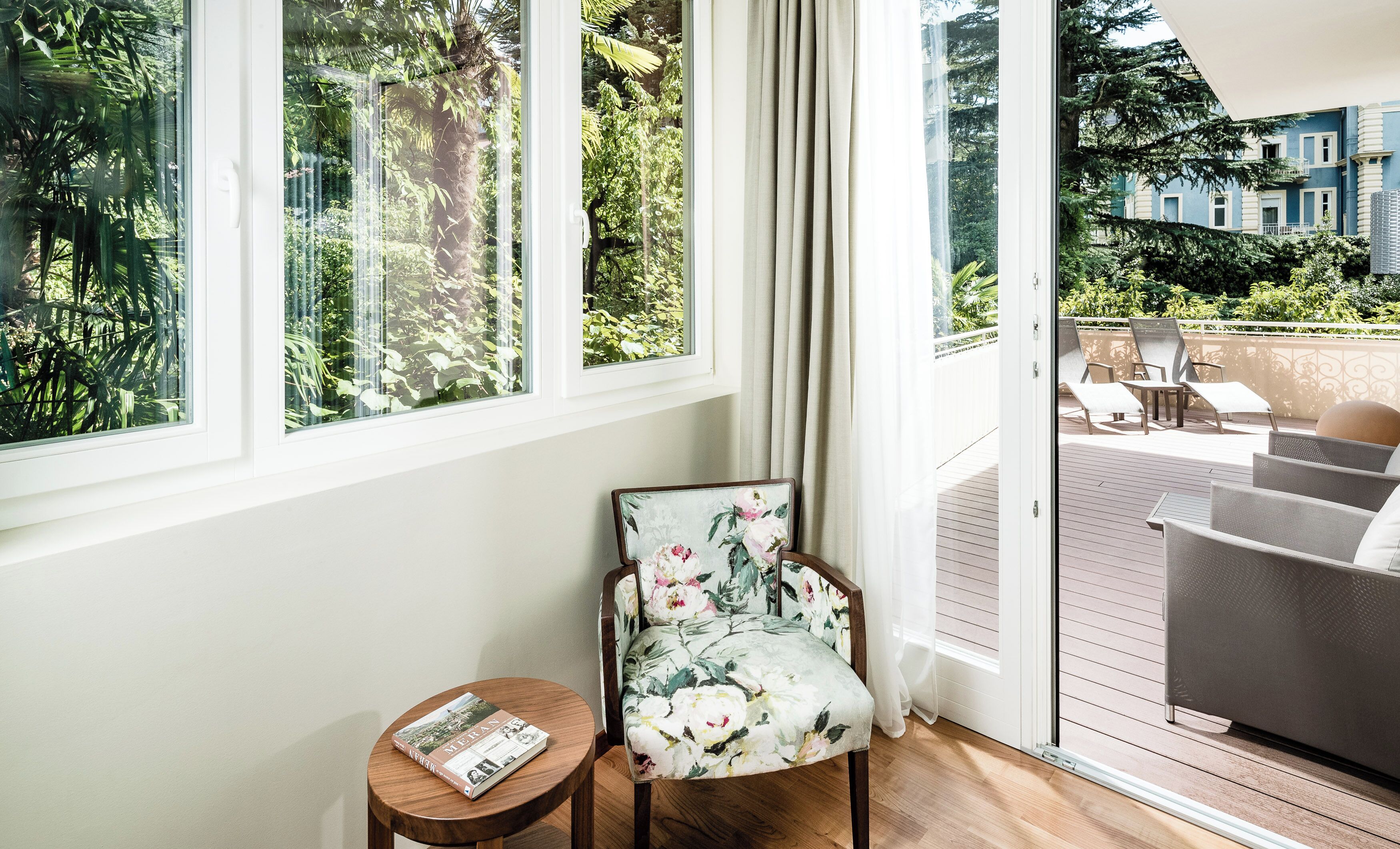 A room with a wooden floor, a chair with a floral pattern, a small round table with a magazine, and a large window with a view of greenery and a building.