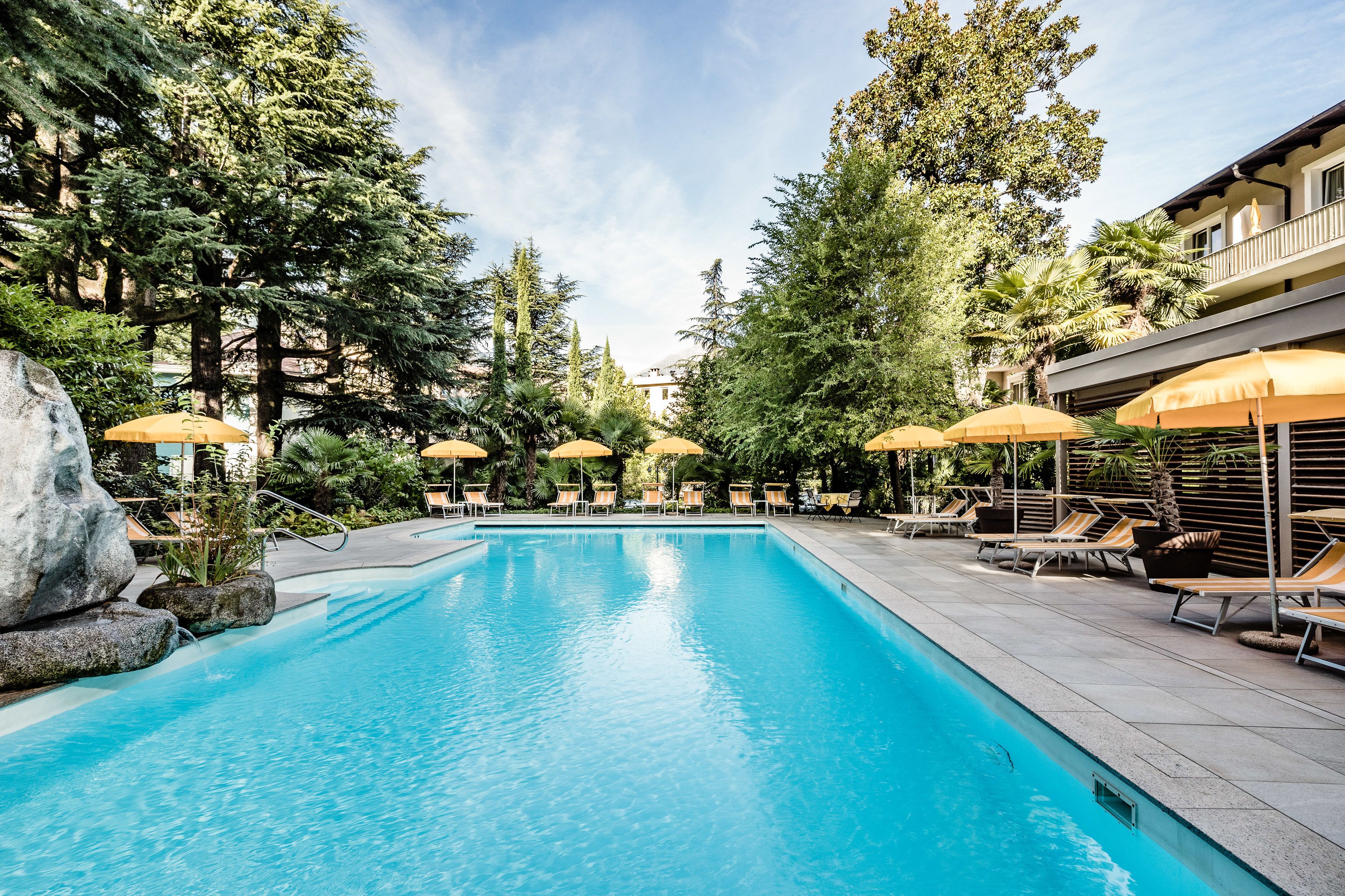 Une piscine entourée d’arbres et d’un bâtiment, avec des chaises longues et des parasols.
