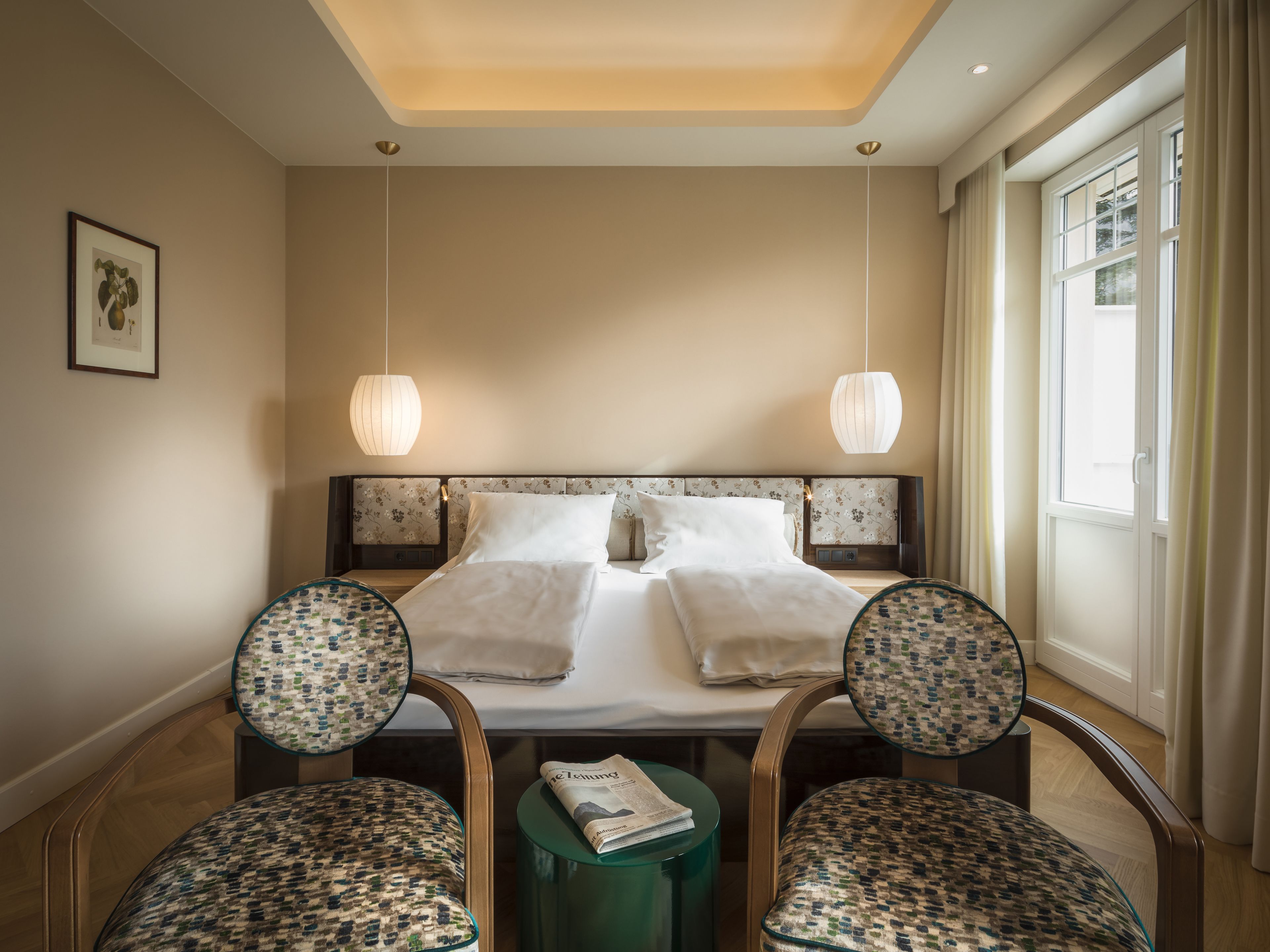 Une chambre d’hôtel avec un grand lit, deux fauteuils rembourrés, une petite table avec un journal et une fenêtre avec des rideaux.