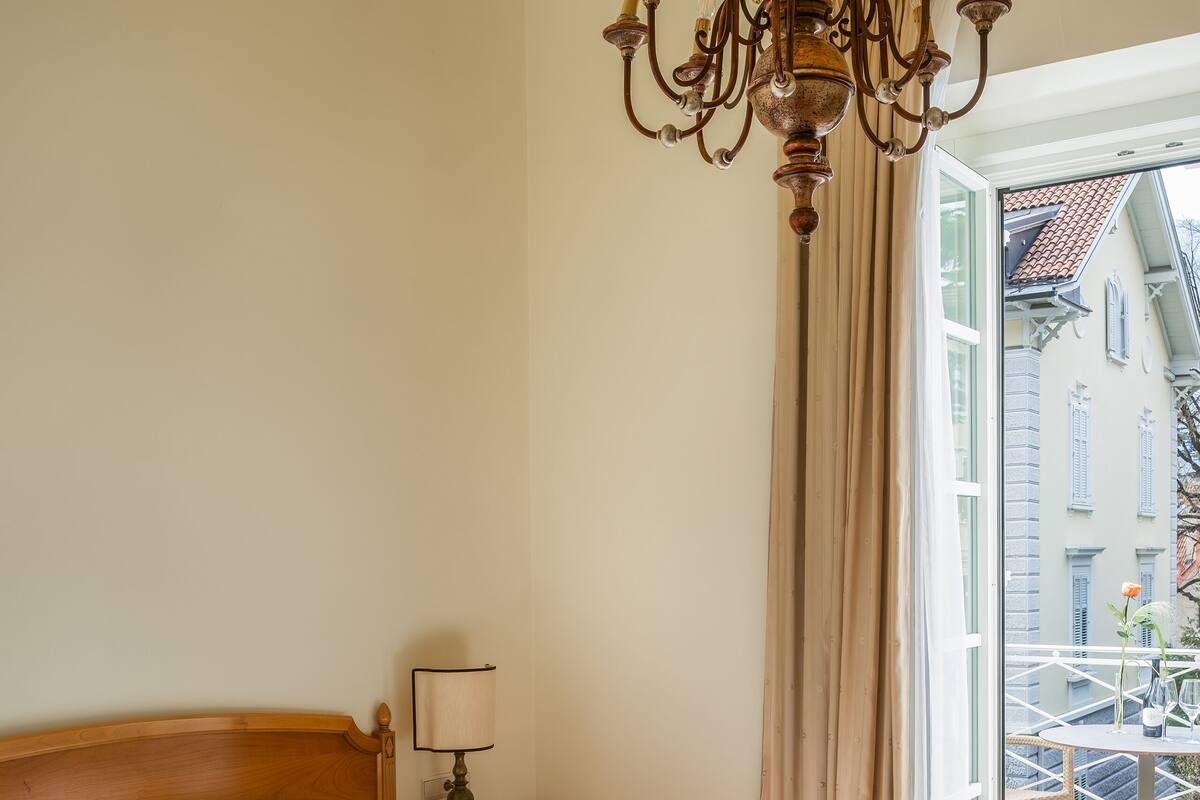 A hotel room with a wooden bed, a chandelier, a nightstand with a lamp, and a view of a balcony with buildings.