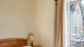 Habitación de hotel con cama de madera, candelabro, mesita de noche con lámpara y vista a un balcón con edificios.