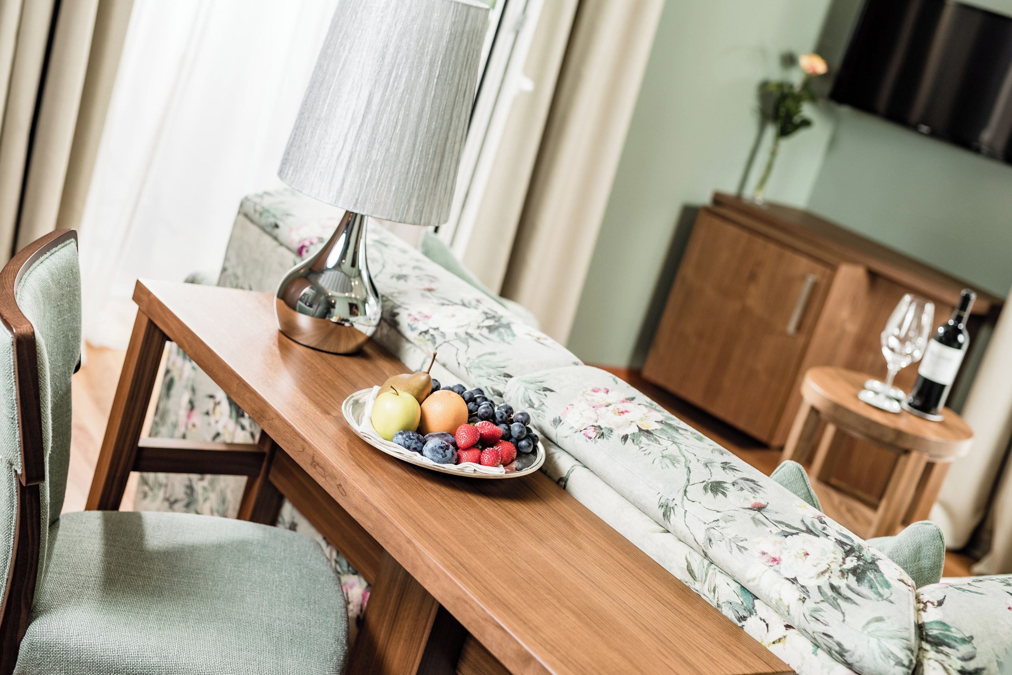 A dining area with a wooden table, floral sofa, and a lamp. A plate of fruit is on the table.