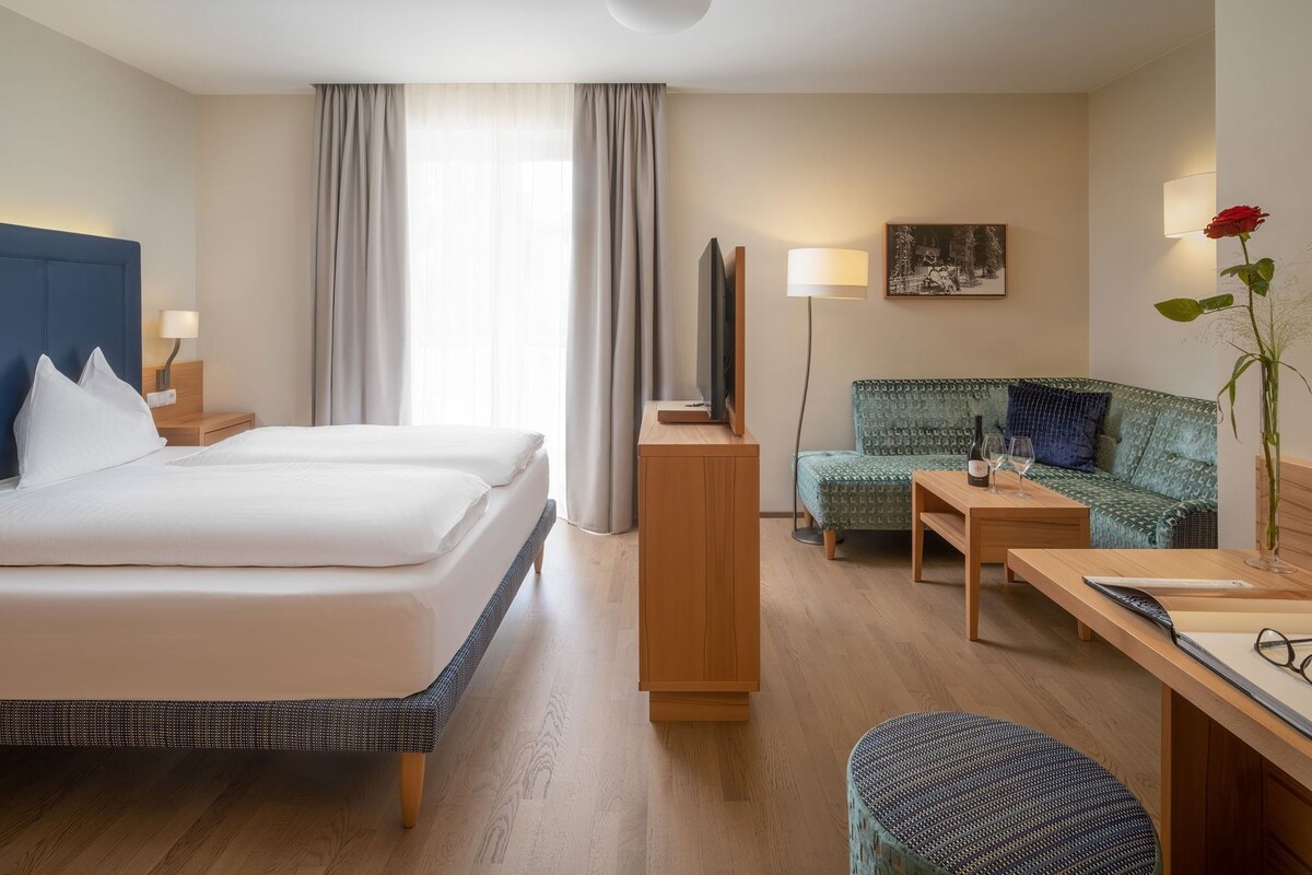 A hotel room with a bed, a desk, a chair, a sofa, a TV, and a painting on the wall.