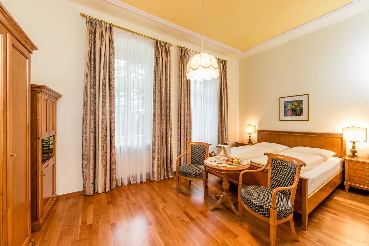 A hotel room with a wooden bed, two chairs, a small table with a fruit plate, a painting on the wall, and a chandelier.