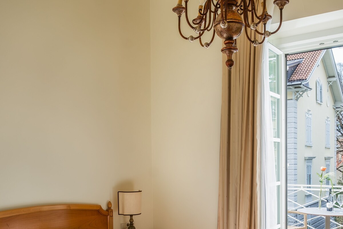 A hotel room with a wooden bed, a chandelier, a nightstand, and a view of the outside.