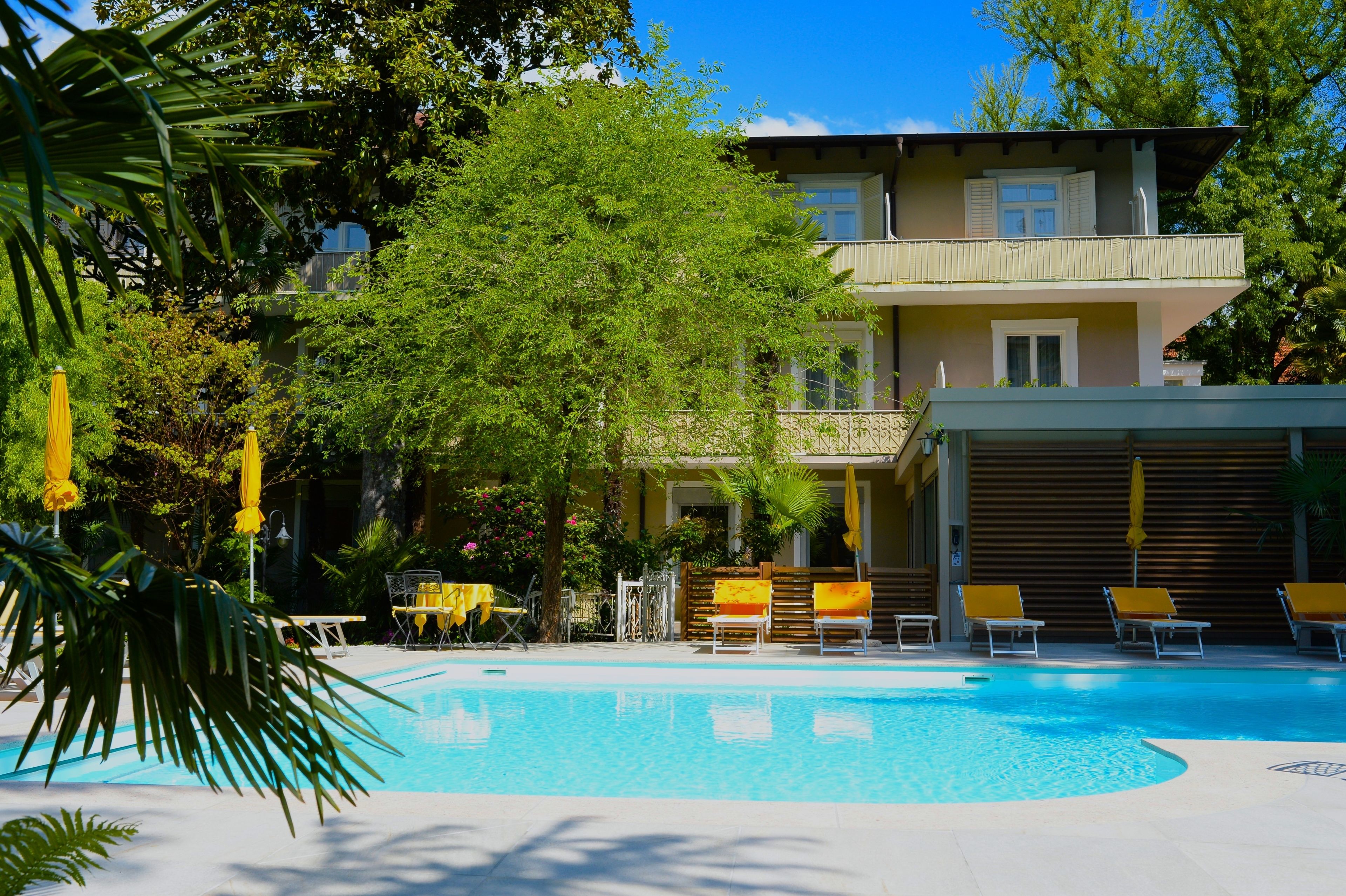 Piscina con sillas de descanso y sombrillas, rodeada de árboles y un edificio con balcones.