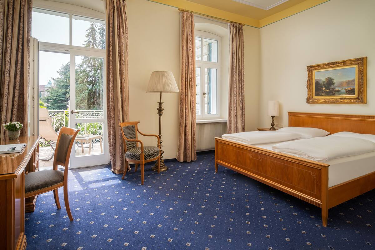 A hotel room with a large bed, a desk with a chair, a lamp, and a window with curtains.