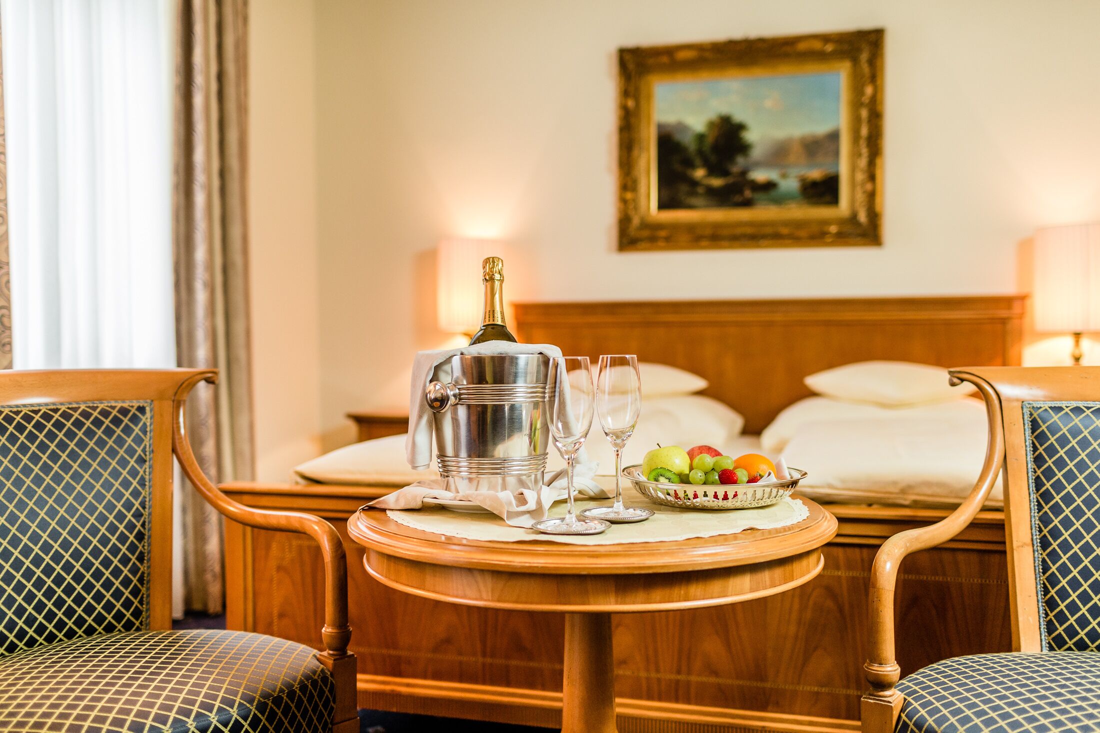 A hotel room with a wooden bed, a bedside table with a bottle, glasses, and a fruit bowl.