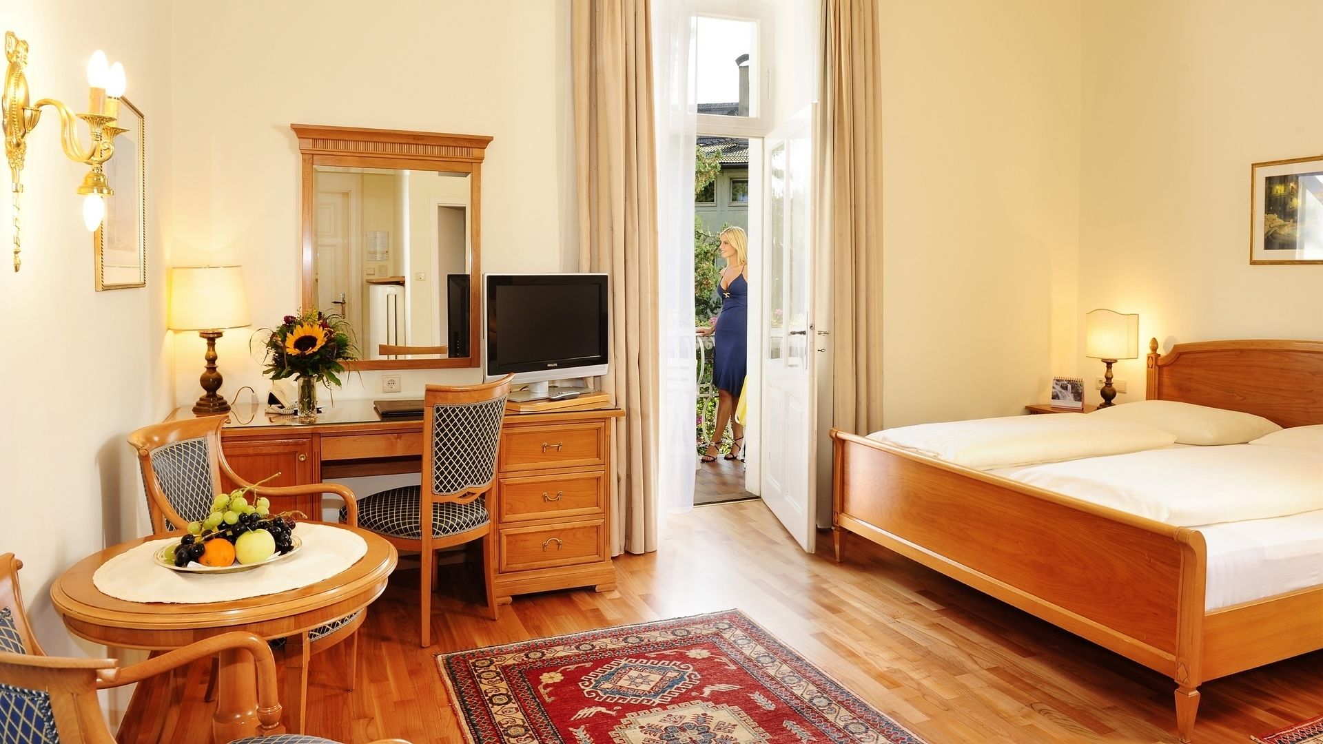 Habitación de hotel con una cama de madera, un escritorio con computadora, una silla, una mesa de comedor con fruta, un espejo y vistas al exterior.