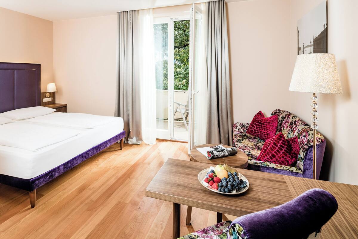 A hotel room with a bed, a table with fruit, a chair, and a view of greenery through the window.