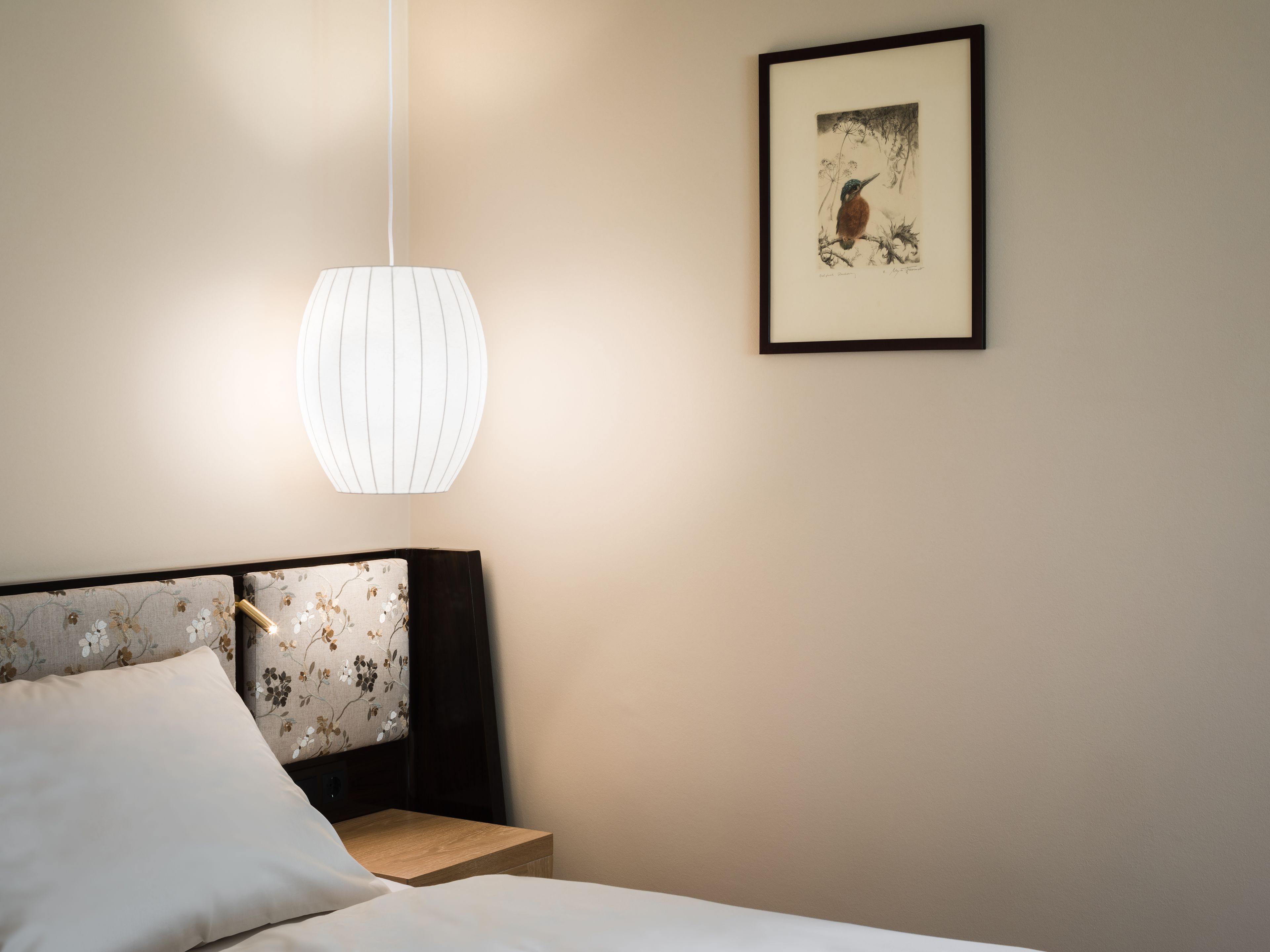 Una cama bien hecha con un cojín, una mesita de noche con una lámpara y un cuadro en la pared.