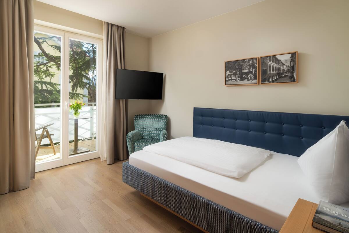A hotel room with a bed, a TV, a chair, and a balcony with a table and plant.
