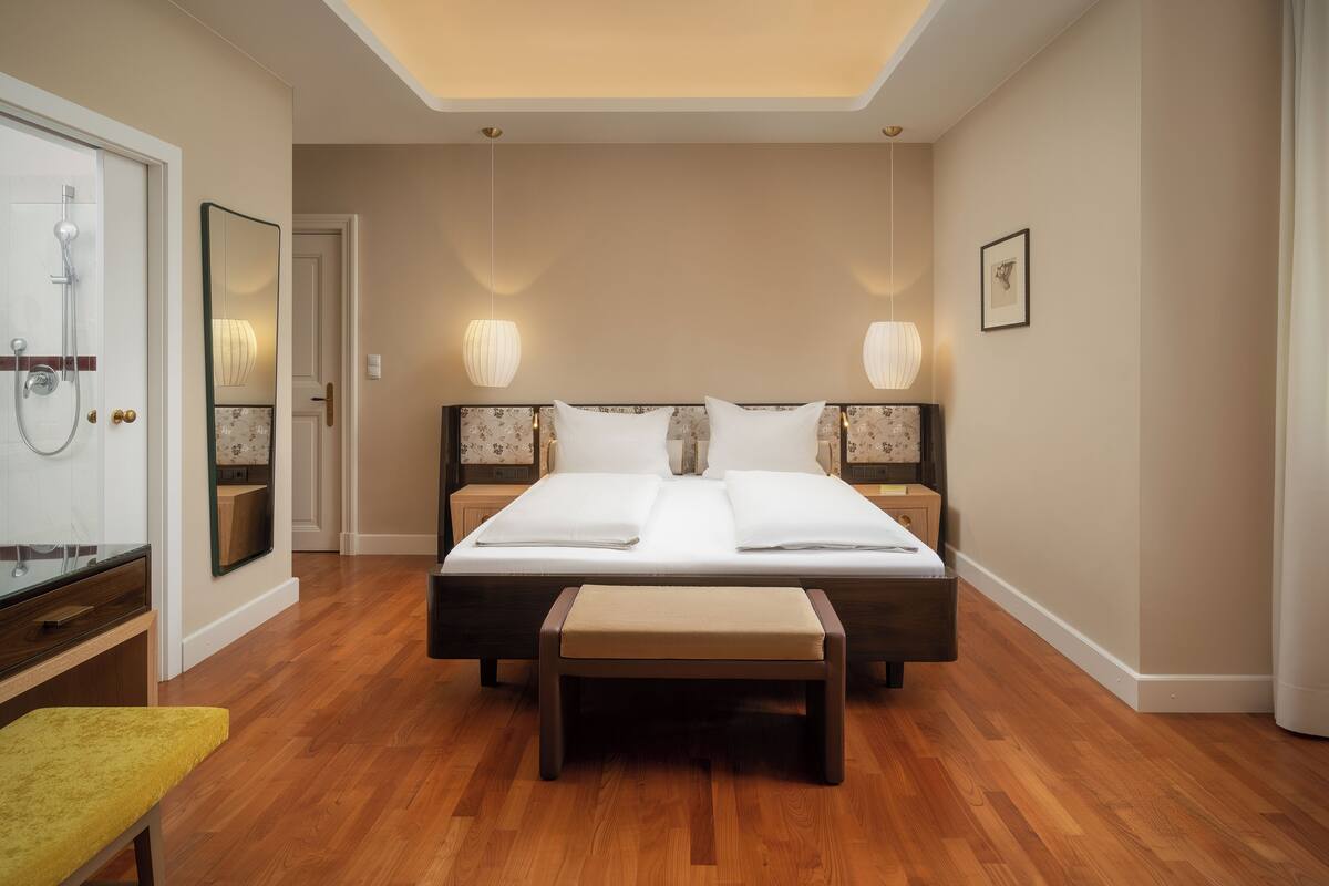 A hotel room with a large bed, bedside lamps, a bench, a mirror, and a shower.