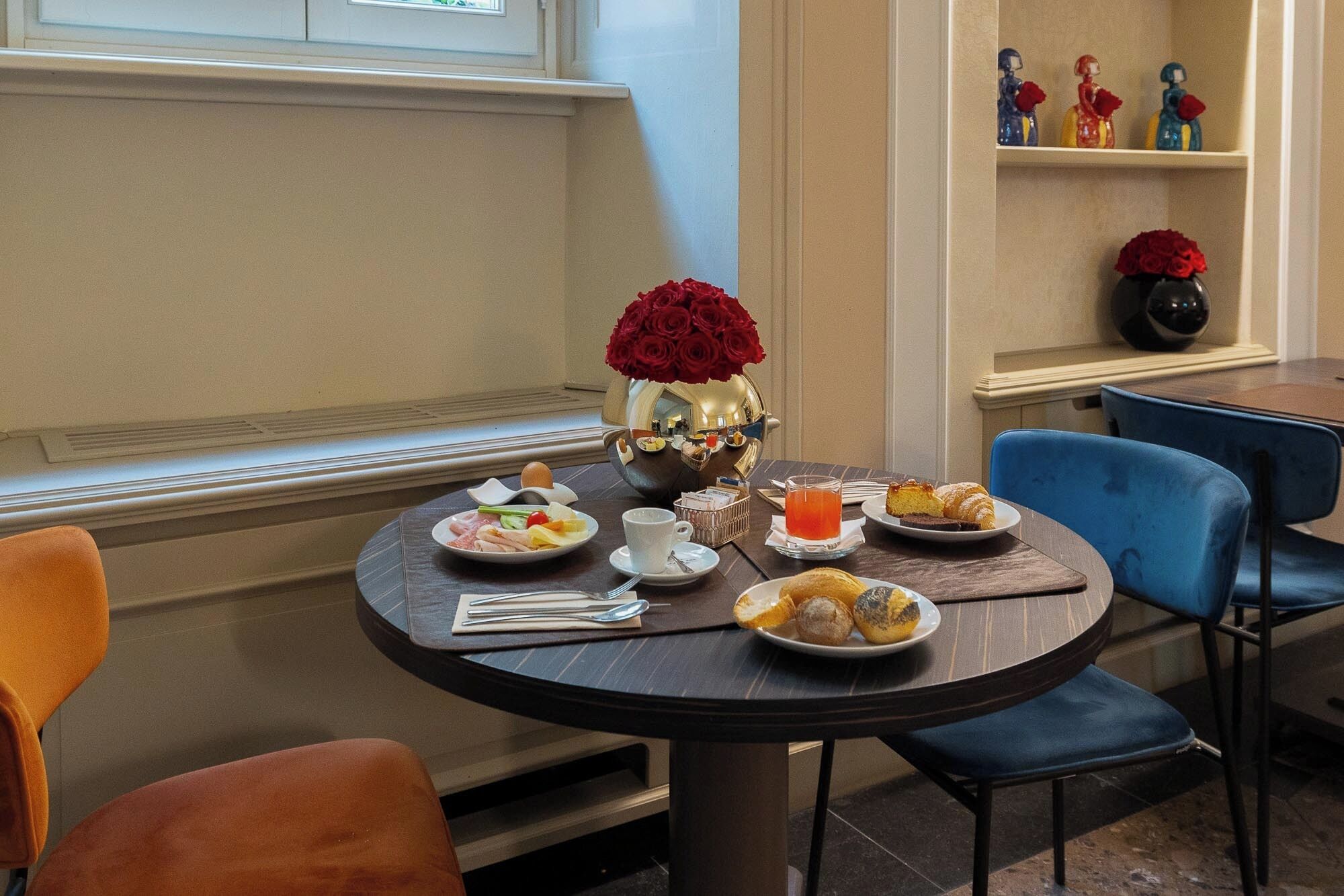 Une table ronde où sont disposés des plats de nourriture, un vase de roses rouges et un verre de jus d’orange.