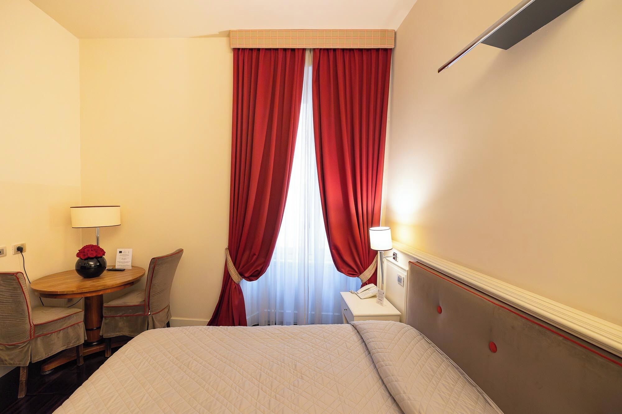 Una habitación de hotel con cama, escritorio con lámpara, silla y ventana con cortinas rojas.