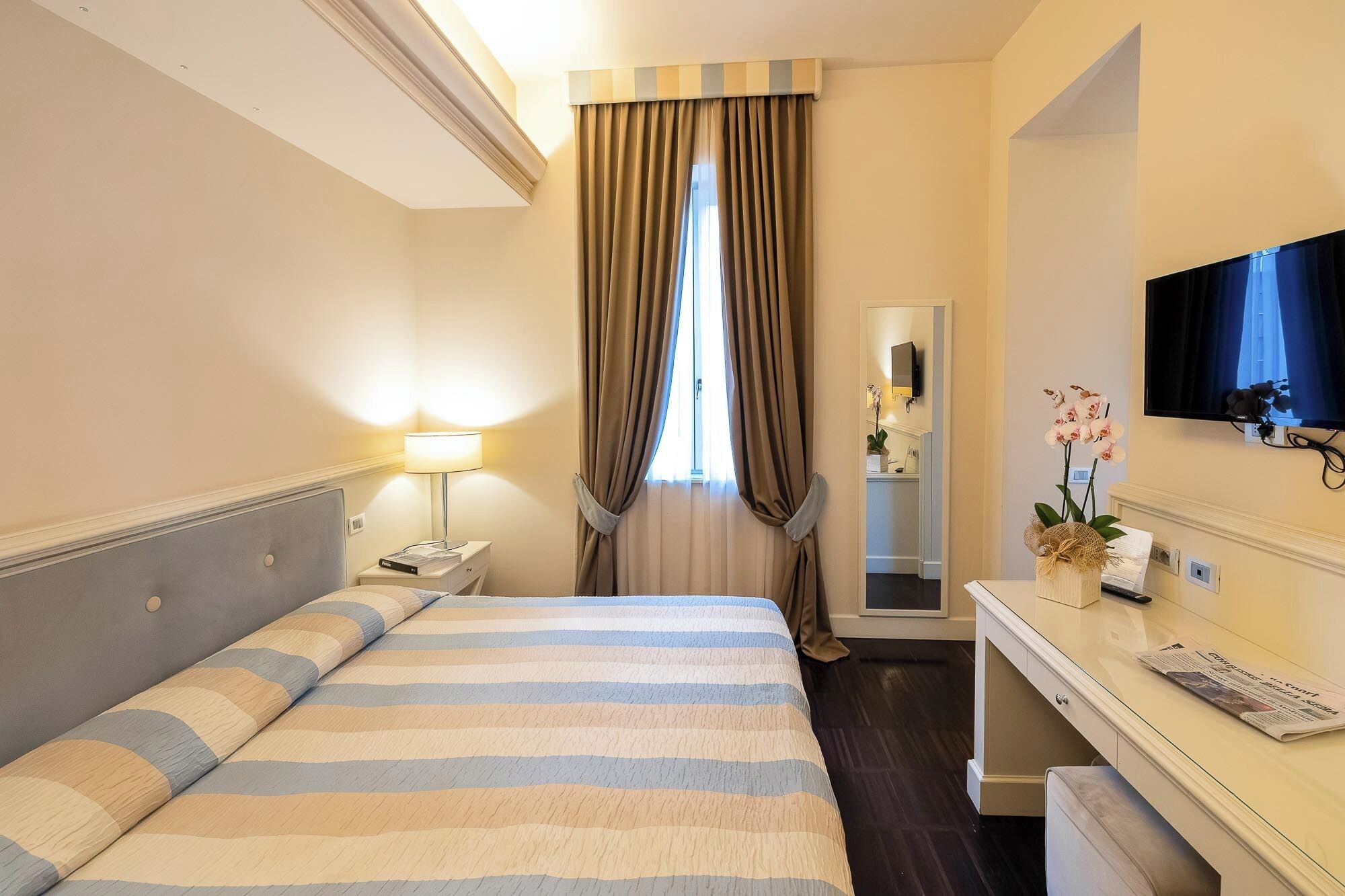 A hotel room with a bed, a desk with a TV, a lamp, and a window with curtains.