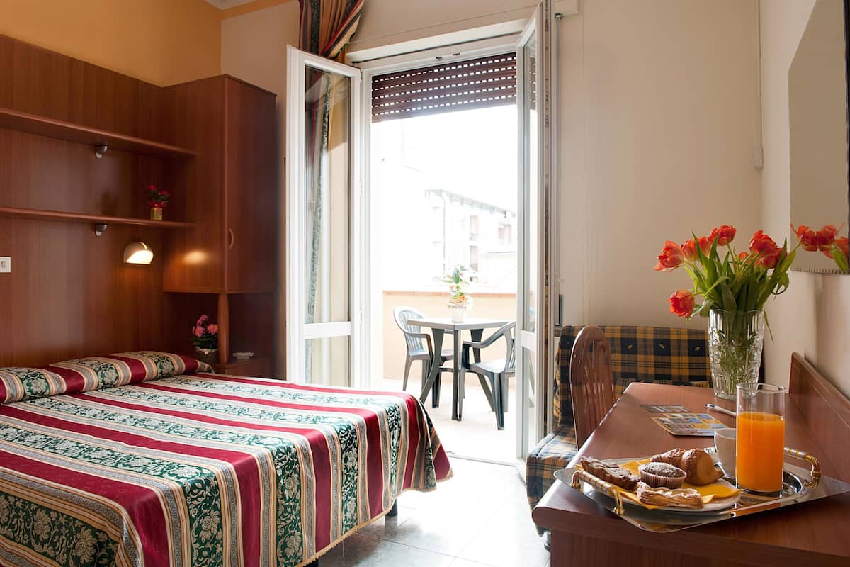 A hotel room with a bed, a dining table with breakfast items, a vase with flowers, and a view of the outside.