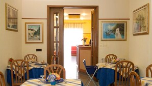 A dining area with tables set for four, each with a blue tablecloth and wooden chairs. There are framed pictures on the walls and a door leading to another room. - Hotel Dolly (Viareggio)