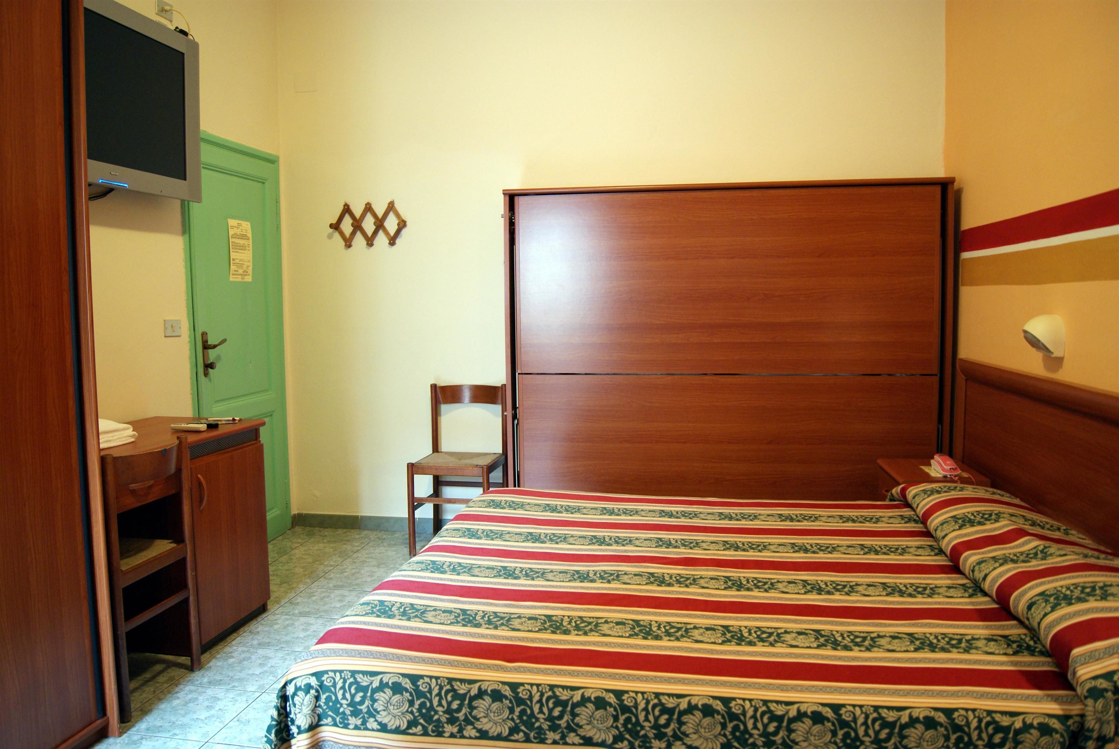 A bedroom with a wooden bed, a desk, a chair, and a television mounted on the wall.