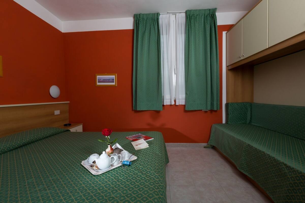 A room with two beds, a wooden headboard, a green patterned bedspread, a green sofa, a window with green curtains, and a small framed picture on the wall.