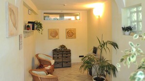 A hallway with a wicker chair, a potted plant, and a wall-mounted shelf with books and decorative items. - Riviera Blu (Pisa)
