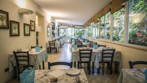 A dining area with tables and chairs, framed pictures on the walls, and large windows with a view of greenery. - Riviera Blu (Pisa)