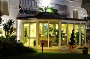 A glass-enclosed building with a roof, surrounded by greenery and potted plants.