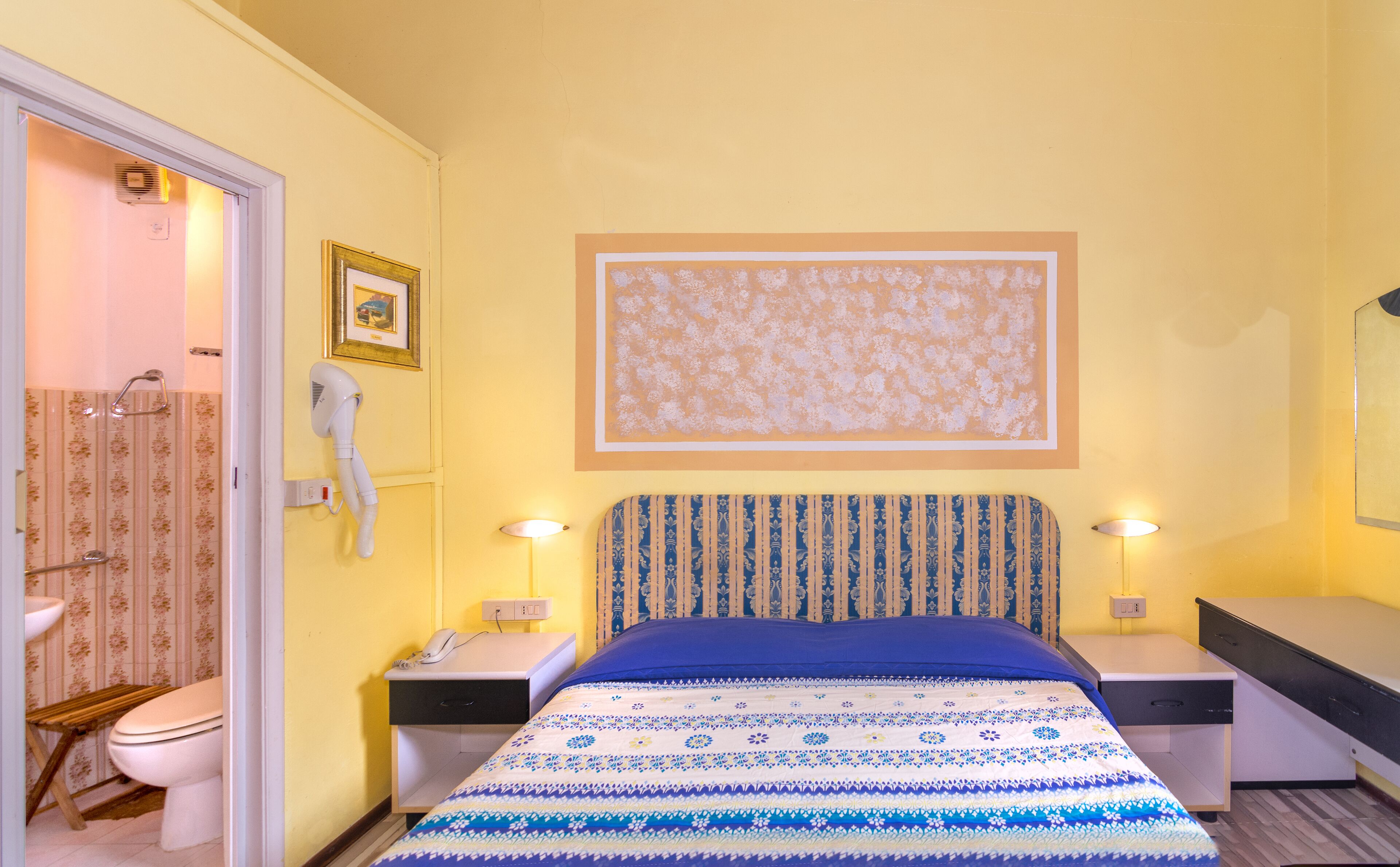 Une chambre d’hôtel comprenant un lit, des tables de chevet, un téléphone, un miroir, un tableau au mur et une salle de bain avec des toilettes et une douche.