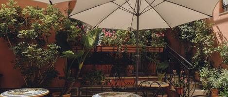An outdoor patio with tables and chairs, surrounded by potted plants and a large umbrella.