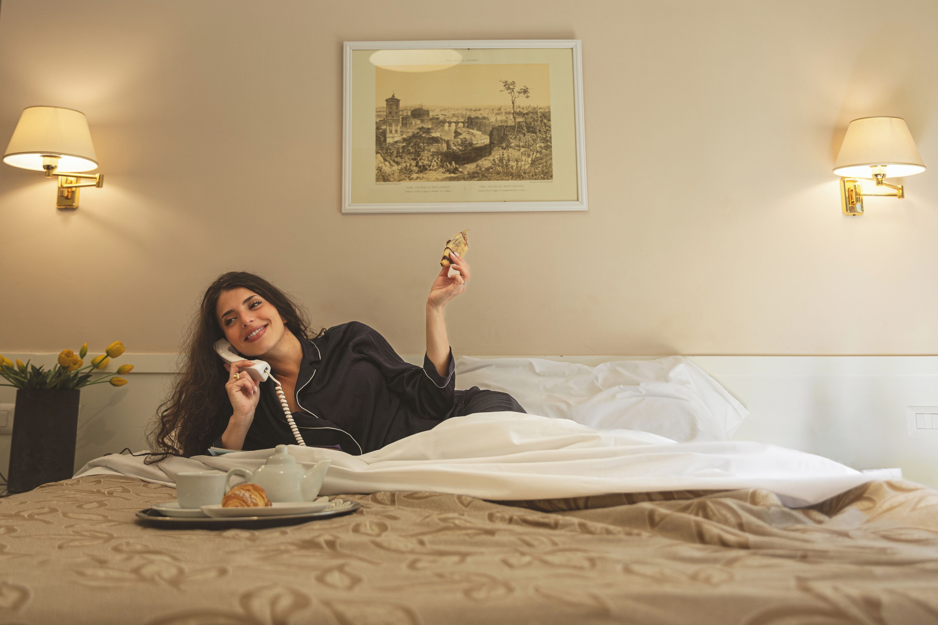 Une personne est allongée sur un lit, avec une tasse et une pâtisserie à portée de main. Un tableau encadré est accroché au mur, et deux lampes sont fixées sur les murs.