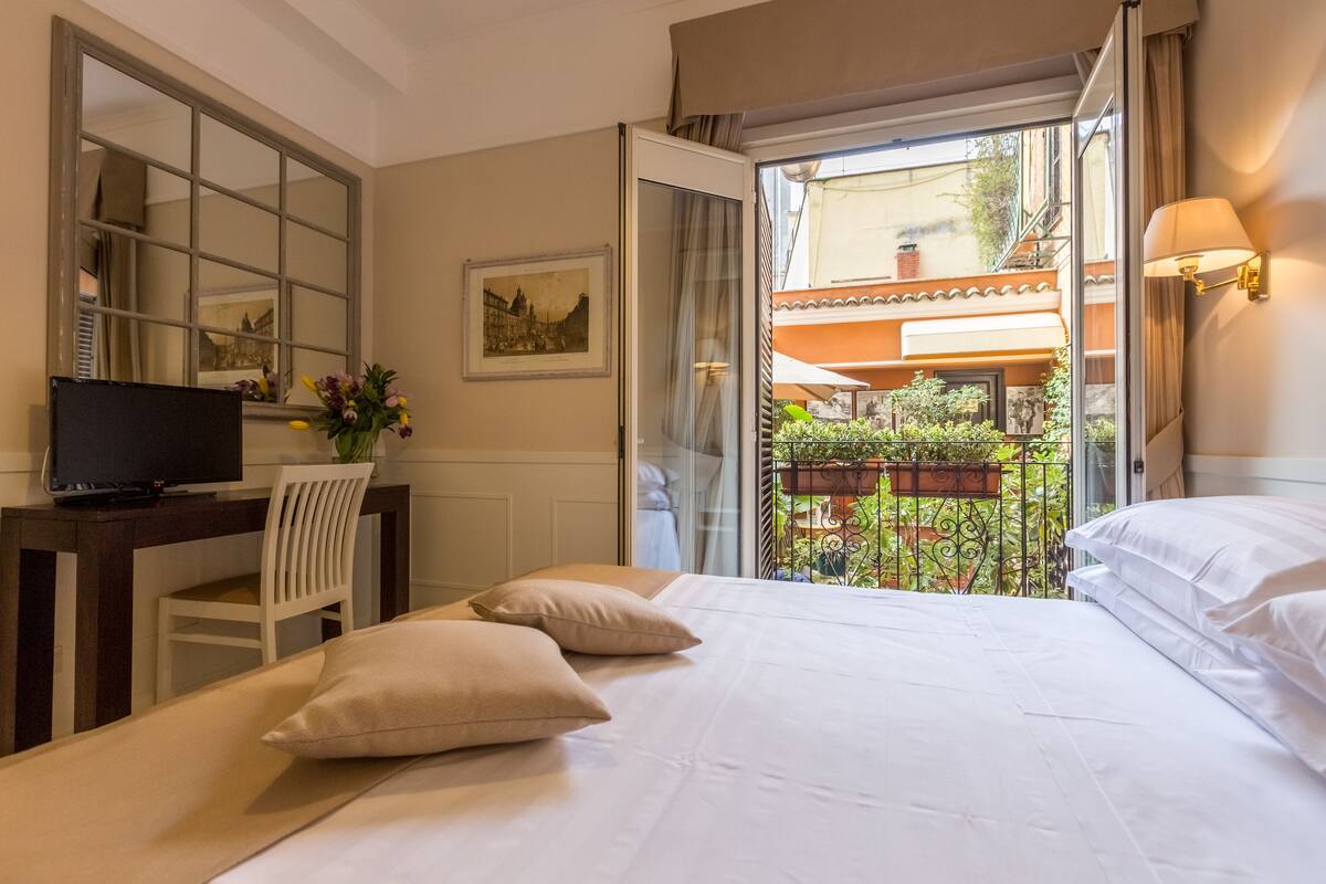 A hotel room with a bed, a desk with a TV, a chair, and a balcony with plants.