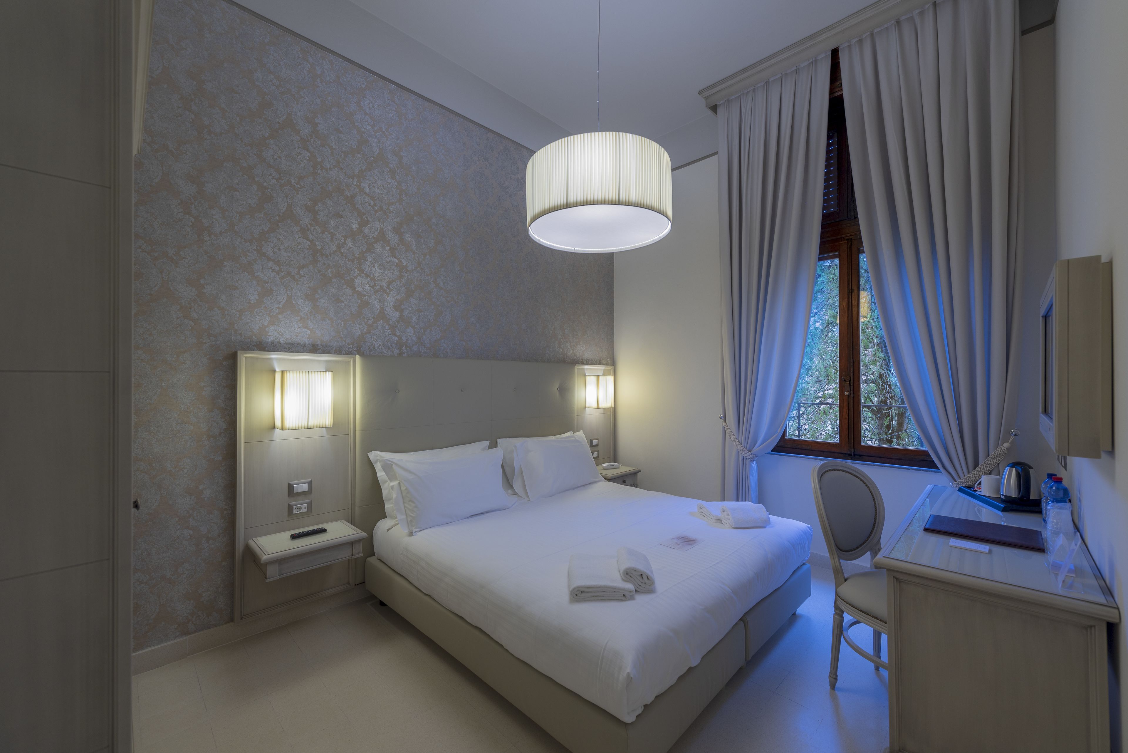 Una habitación de hotel con cama, lámparas de noche, escritorio y ventana con cortinas.