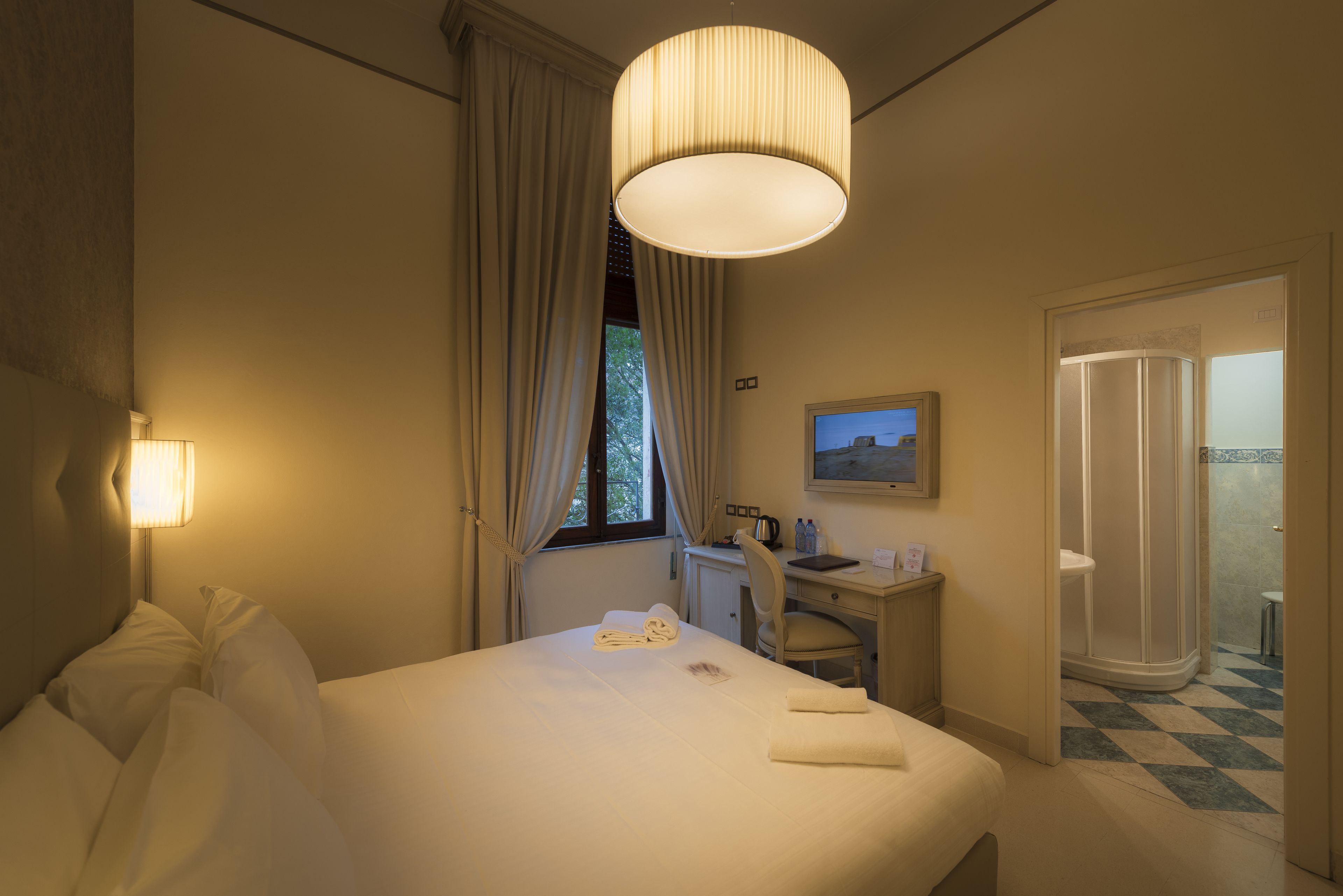 Quarto de hotel com cama, escrivaninha, cadeira, luminária de mesa, janela com cortinas, um quadro na parede e banheiro com chão quadriculado.