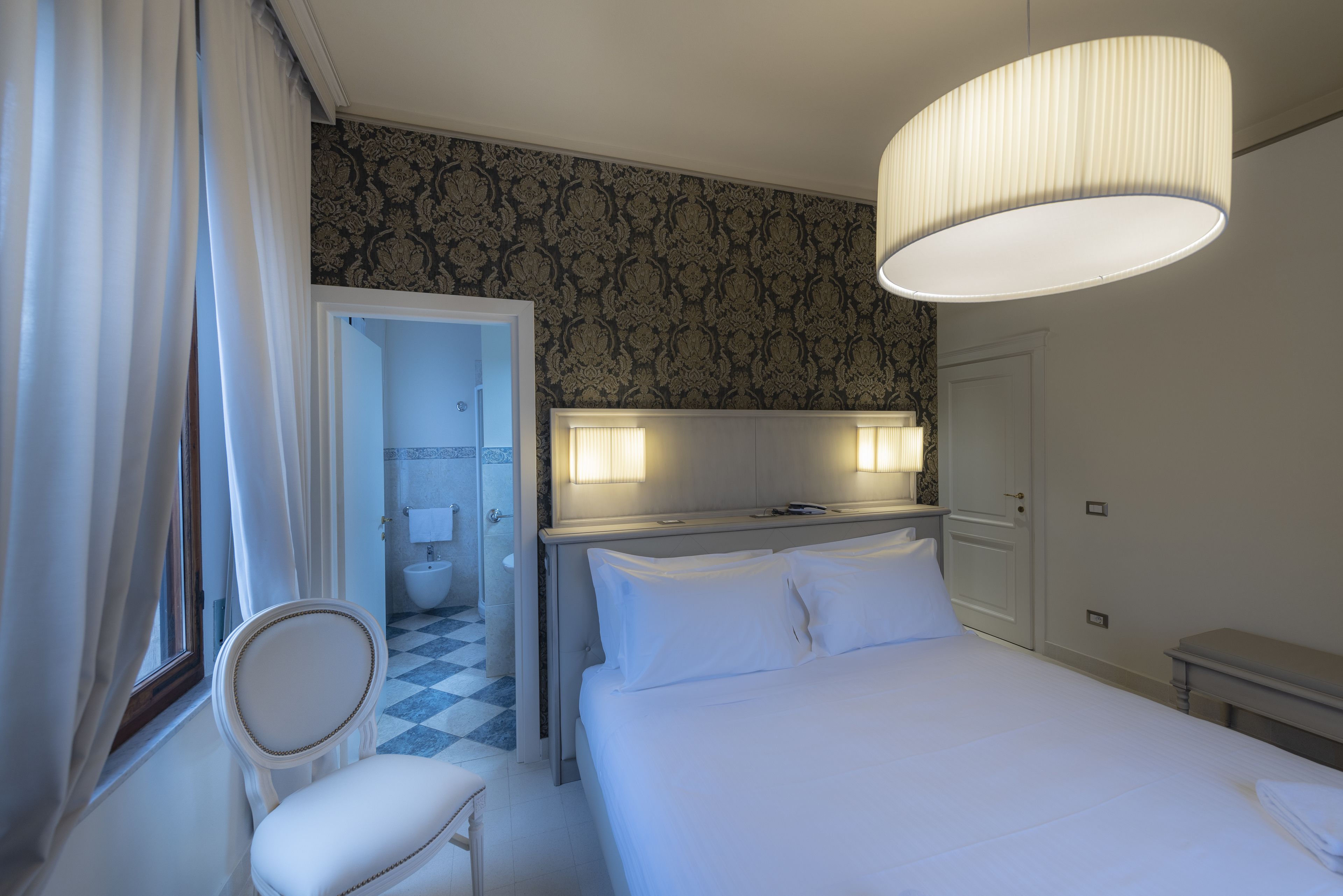 Ein Hotelzimmer mit Bett, Stuhl, Schreibtisch, Spiegel und einem durch eine offene Tür einsehbaren Badezimmer.