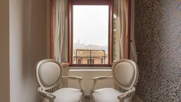 A room with two white upholstered chairs facing a window with curtains, a radiator below, and patterned wallpaper on the right wall.