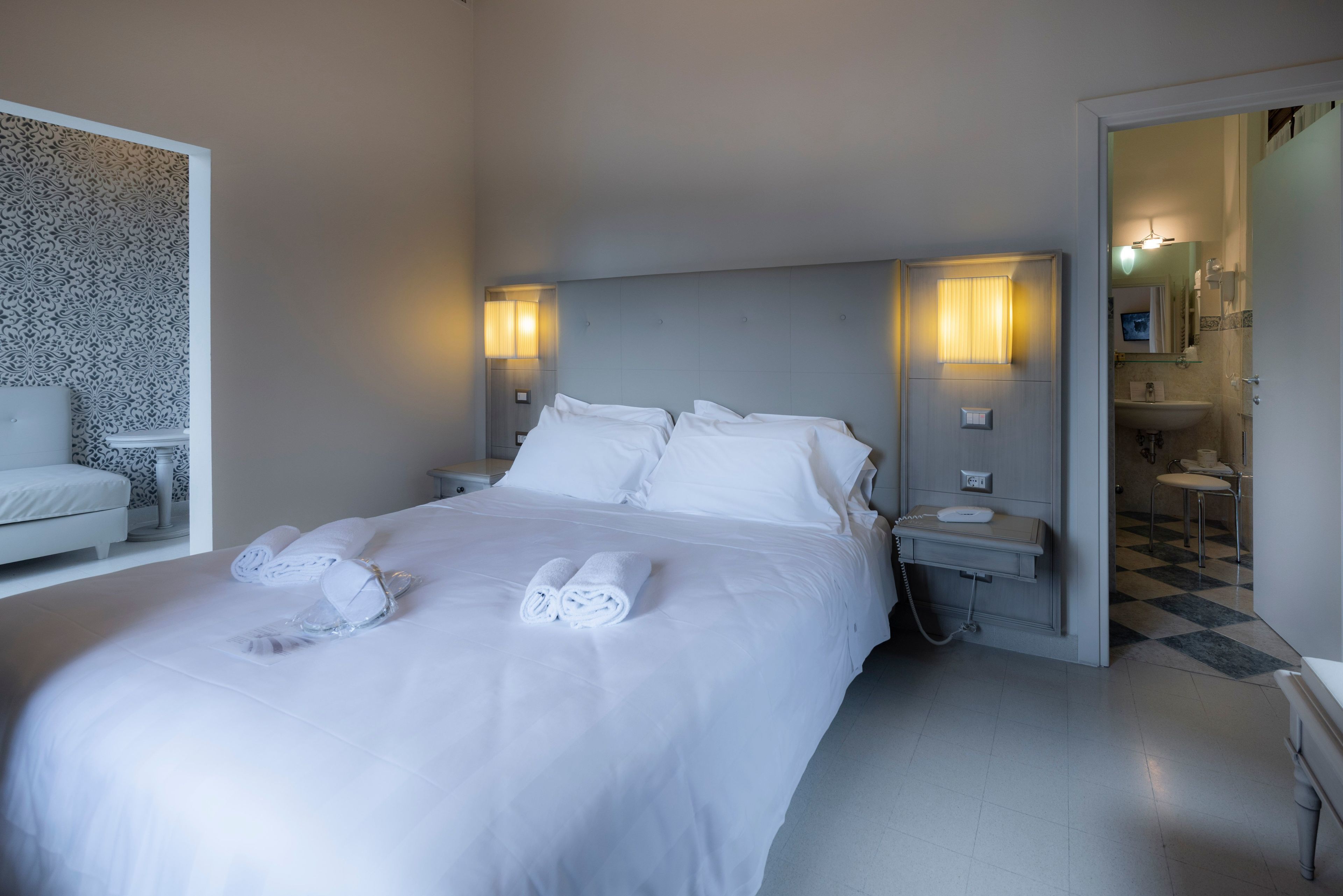 Ein Hotelzimmer mit einem großen Bett, zwei Nachttischlampen, einem Nachttisch, einem durchgegangene Tür erkennbaren Badezimmer und einem Sessel in der Ecke.