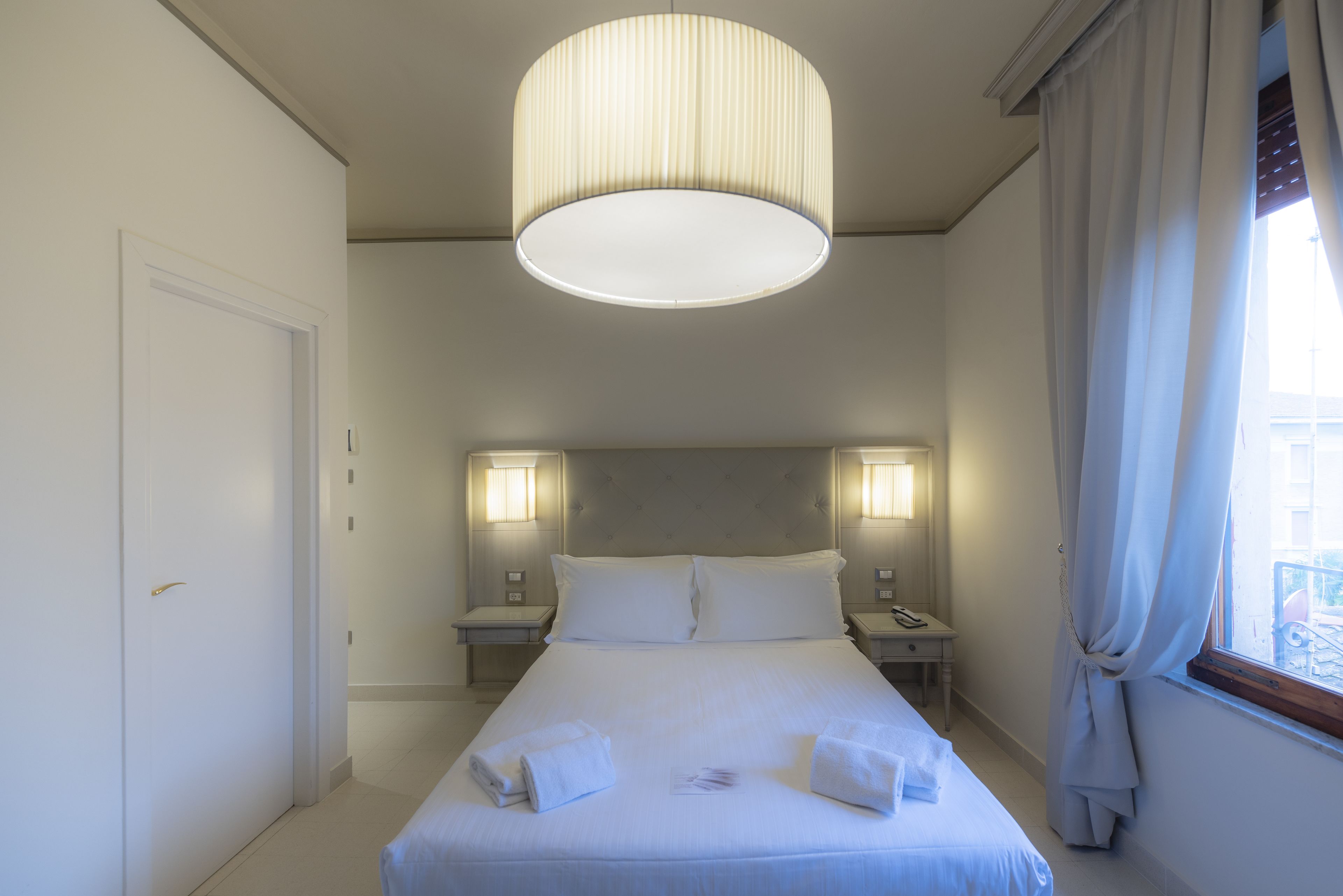 A hotel room with a large bed, two bedside tables, a nightstand, a ceiling light, and a window with curtains.