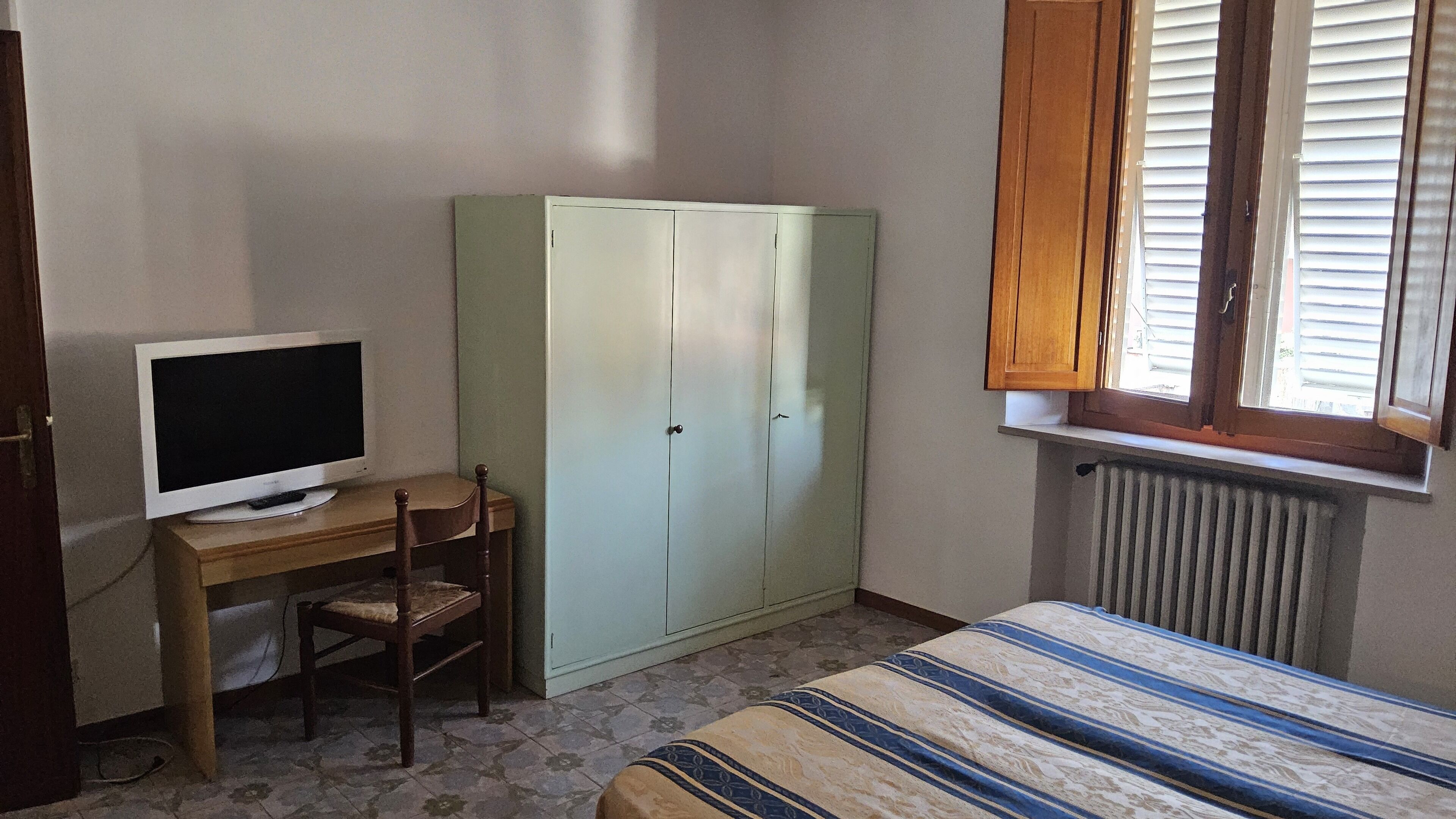 Um quarto com uma cama, uma escrivaninha com computador, um guarda-roupa e uma janela com persianas.