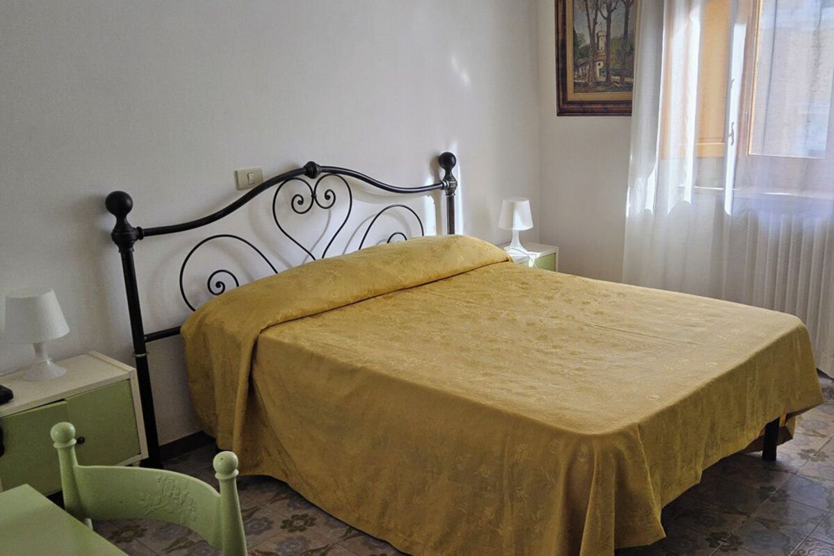 Una cama individual con una colcha amarilla en una habitación con pared blanca y ventana con cortinas de gasa.