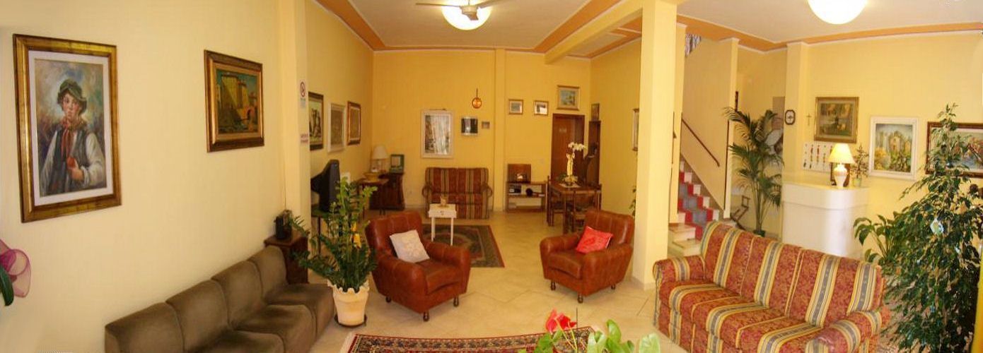 Uma sala de estar com sofá, poltronas e uma mesa de centro. Há quadros nas paredes e uma escada ao fundo.
